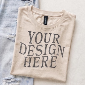 May include: A beige crew neck t-shirt with the text YOUR DESIGN HERE printed in a distressed dark gray font on the front. The shirt is positioned next to a pair of light blue distressed denim jeans with visible rips and frayed edges.