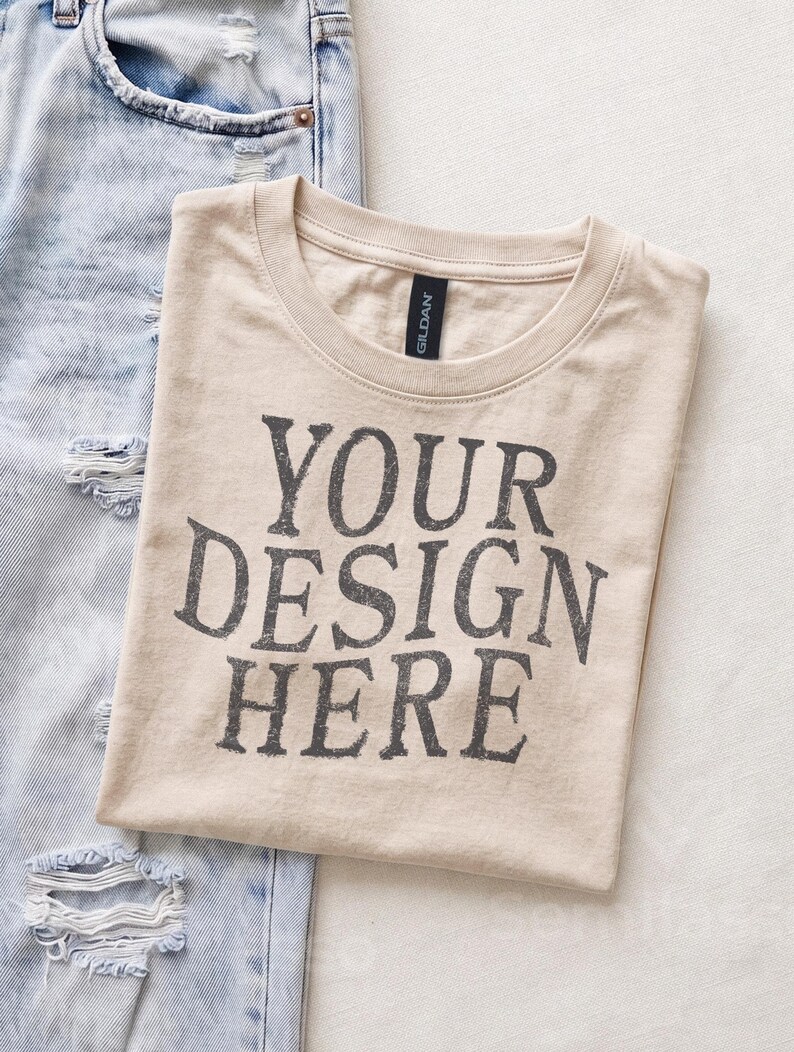May include: A beige crew neck t-shirt with the text YOUR DESIGN HERE printed in a distressed dark gray font on the front. The shirt is positioned next to a pair of light blue distressed denim jeans with visible rips and frayed edges.