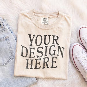 May include: A beige short-sleeved t-shirt with a crew neckline and a Comfort Colors brand label at the collar. The front of the shirt features the black text "YOUR DESIGN HERE" in a bold, distressed, serif font. The shirt is size medium.