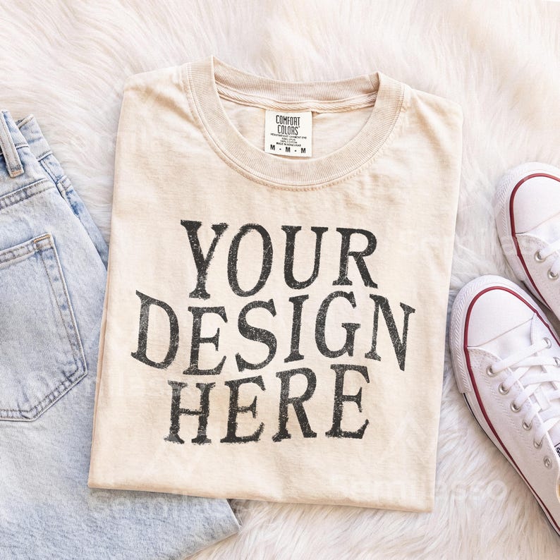 May include: A beige short-sleeved t-shirt with a crew neckline and a Comfort Colors brand label at the collar. The front of the shirt features the black text "YOUR DESIGN HERE" in a bold, distressed, serif font. The shirt is size medium.