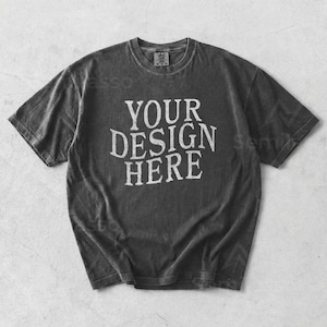 Puede incluir: Camiseta gris oscuro con el texto blanco "YOUR DESIGN HERE". La camiseta tiene cuello redondo y mangas cortas. La tela tiene un aspecto descolorido y vintage. Es probable que la camiseta esté pre-encogida.