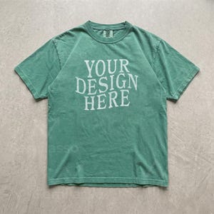May include: A short-sleeved crew neck t-shirt in a muted sage green color. The front of the garment features the text "YOUR DESIGN HERE" printed in a distressed, off-white, serif font. The shirt has a standard fit with a ribbed collar neckline.