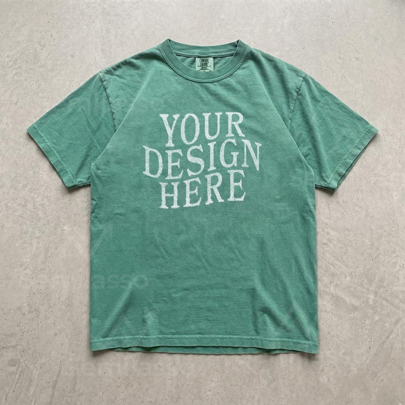 May include: A short-sleeved crew neck t-shirt in a muted sage green color. The front of the garment features the text "YOUR DESIGN HERE" printed in a distressed, off-white, serif font. The shirt has a standard fit with a ribbed collar neckline.