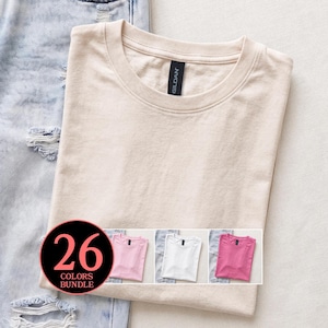 Könnte beinhalten: Ein beiges T-Shirt mit Rundhalsausschnitt und Gildan-Markenetikett am Innenkragen, platziert neben einer hellblauen, abgenutzten Jeans. Eine kreisförmige Grafik zeigt den Text 26 COLORS BUNDLE mit drei kleineren T-Shirt-Farboptionen.