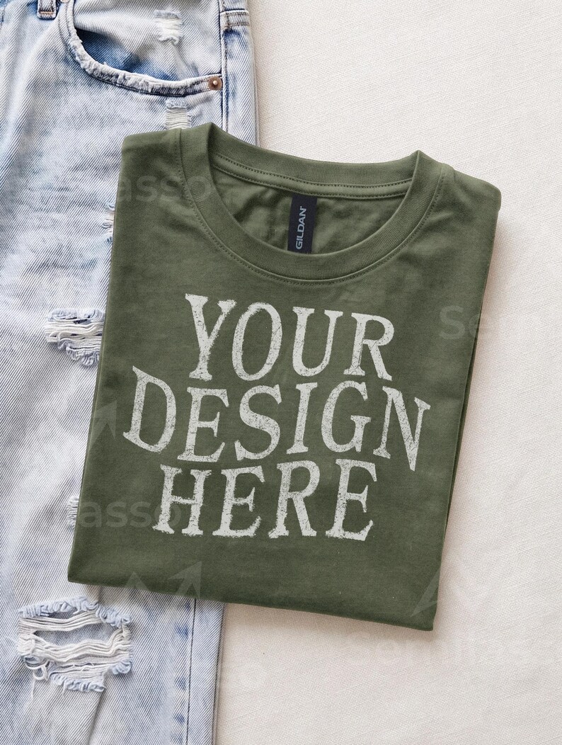 May include: An olive green crew neck t-shirt with the white text YOUR DESIGN HERE printed on the front. The shirt features a Gildan brand label at the inner neckline and is positioned next to a pair of light blue distressed denim jeans.