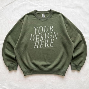Puede incluir: Una sudadera de cuello redondo en color verde oliva con mangas largas y bordes acanalados en el cuello, pu&ntilde;os y dobladillo. En el centro del pecho aparece el texto "YOUR DESIGN HERE" en una fuente serif desgastada de color blanco roto. Etiqueta Gildan.