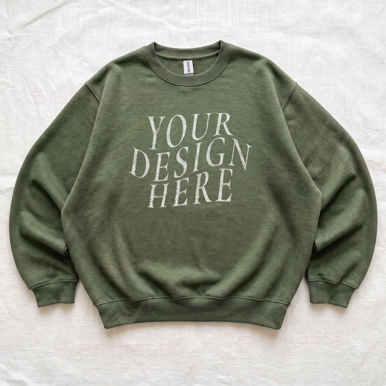 Puede incluir: Una sudadera de cuello redondo en color verde oliva con mangas largas y bordes acanalados en el cuello, pu&ntilde;os y dobladillo. En el centro del pecho aparece el texto "YOUR DESIGN HERE" en una fuente serif desgastada de color blanco roto. Etiqueta Gildan.