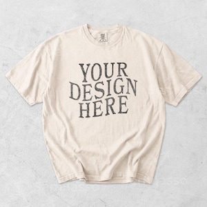 May include: Cream-colored t-shirt with the text "YOUR DESIGN HERE" in a distressed, dark gray font. The shirt has a crew neck and short sleeves, and is likely made of cotton or a similar fabric.