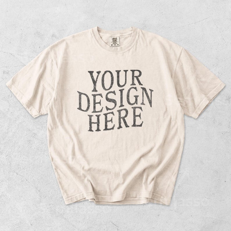May include: Cream-colored t-shirt with the text "YOUR DESIGN HERE" in a distressed, dark gray font. The shirt has a crew neck and short sleeves, and is likely made of cotton or a similar fabric.