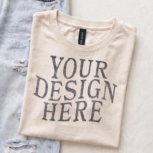 May include: A beige crew neck t-shirt with the black text YOUR DESIGN HERE printed on the front. The shirt features a Gildan brand label at the inner neckline and is positioned next to a pair of light blue distressed denim jeans with frayed holes.