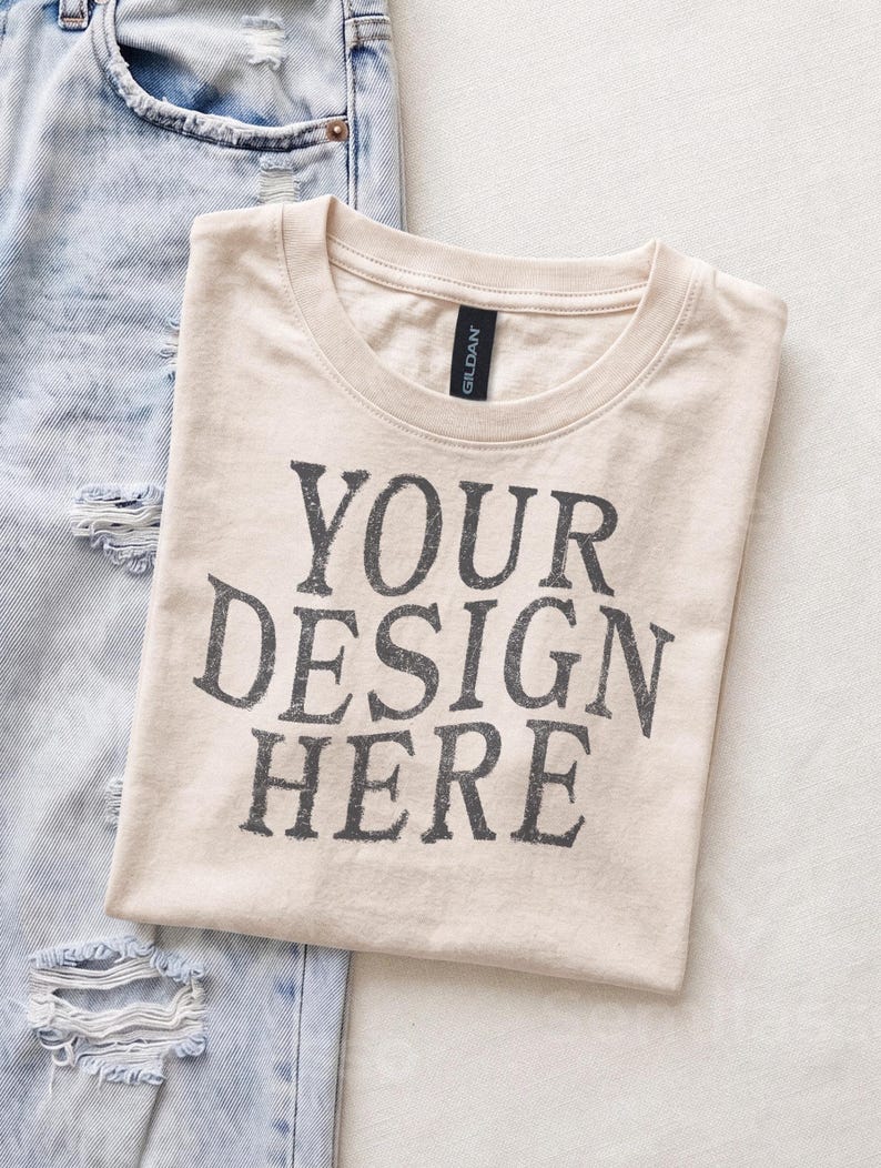 May include: A beige crew neck t-shirt with the black text YOUR DESIGN HERE printed on the front. The shirt features a Gildan brand label at the inner neckline and is positioned next to a pair of light blue distressed denim jeans with frayed holes.