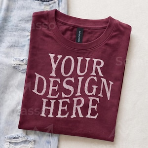 May include: A maroon crew neck t-shirt with the white distressed text YOUR DESIGN HERE printed on the front. The shirt features a black Gildan brand label at the inner neckline and is positioned next to a pair of light blue distressed denim jeans.