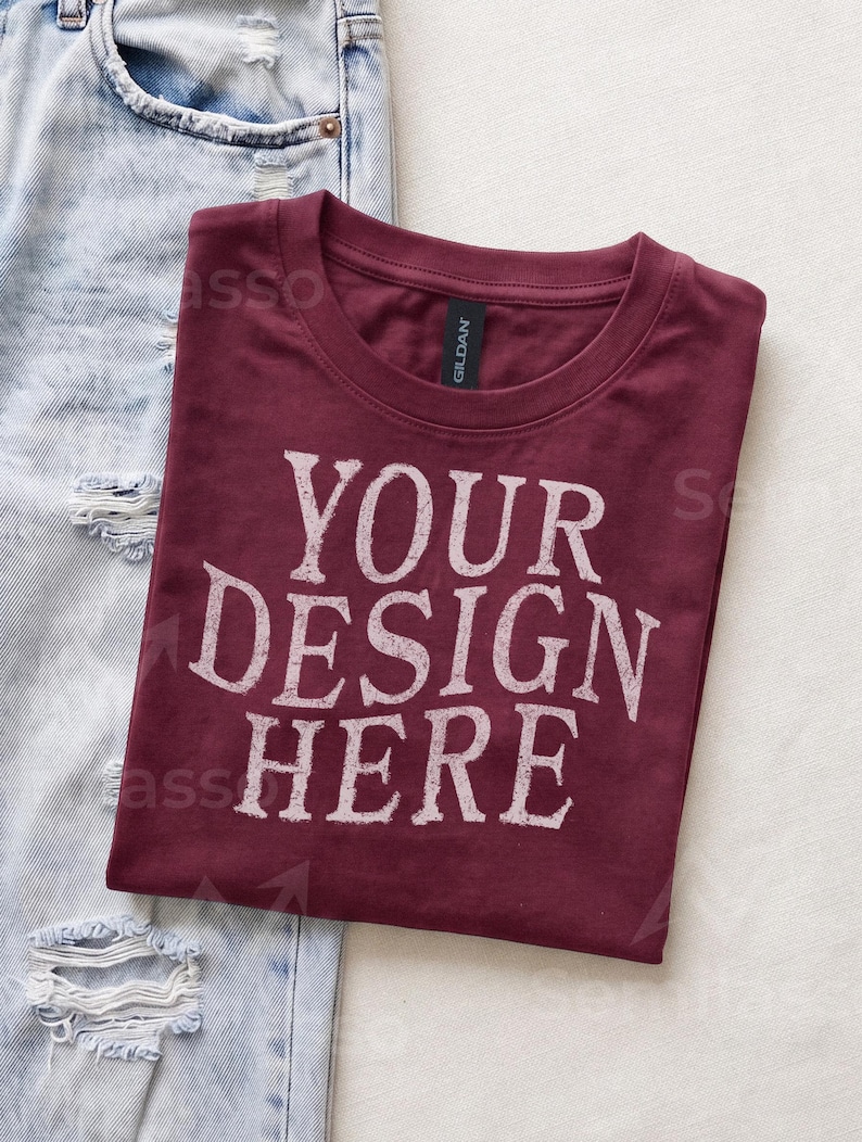 May include: A maroon crew neck t-shirt with the white distressed text YOUR DESIGN HERE printed on the front. The shirt features a black Gildan brand label at the inner neckline and is positioned next to a pair of light blue distressed denim jeans.