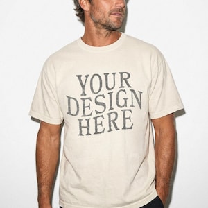 Puede incluir: Camiseta color crema con el texto "YOUR DESIGN HERE" en una fuente gris desgastada. La camiseta de manga corta está hecha de un material suave y tiene un cuello redondo clásico.