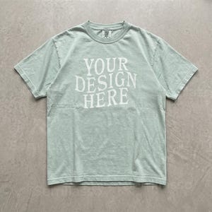 May include: A light sage green short-sleeve crew neck t-shirt featuring the white text "YOUR DESIGN HERE" printed in a bold, serif font across the center of the chest. The garment has a standard fit with a ribbed collar and visible stitching.