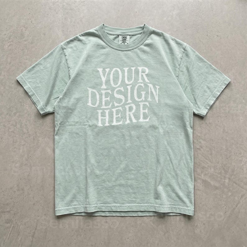May include: A light sage green short-sleeve crew neck t-shirt featuring the white text "YOUR DESIGN HERE" printed in a bold, serif font across the center of the chest. The garment has a standard fit with a ribbed collar and visible stitching.