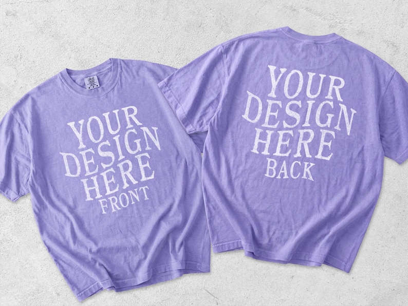May include: Two light purple short-sleeve t-shirts displayed together. The left shirt features the white text YOUR DESIGN HERE FRONT. The right shirt shows the back side with the white text YOUR DESIGN HERE BACK. Both shirts have a crew neckline.