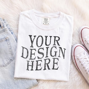 May include: A white short-sleeved t-shirt with a crew neckline featuring the black distressed text YOUR DESIGN HERE printed on the front. The shirt has a Comfort Colors brand label at the inner neck and is displayed alongside light blue denim jeans.
