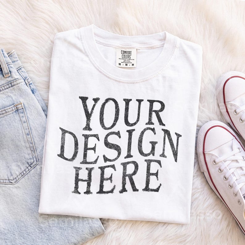 May include: A white short-sleeved t-shirt with a crew neckline featuring the black distressed text YOUR DESIGN HERE printed on the front. The shirt has a Comfort Colors brand label at the inner neck and is displayed alongside light blue denim jeans.