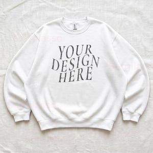 Puede incluir: Una sudadera blanca de cuello redondo con el texto negro "YOUR DESIGN HERE" impreso en una fuente serif desgastada en el centro del pecho. La prenda tiene mangas largas con pu&ntilde;os y dobladillo acanalados, presentando un estilo cl&aacute;sico.
