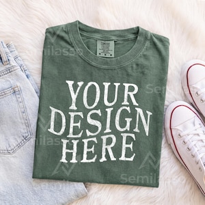 May include: A dark olive green crew neck t-shirt featuring the white text "YOUR DESIGN HERE" printed in a distressed, bold font on the front. The shirt has a visible interior neck label indicating a size medium and a classic short-sleeve construction.