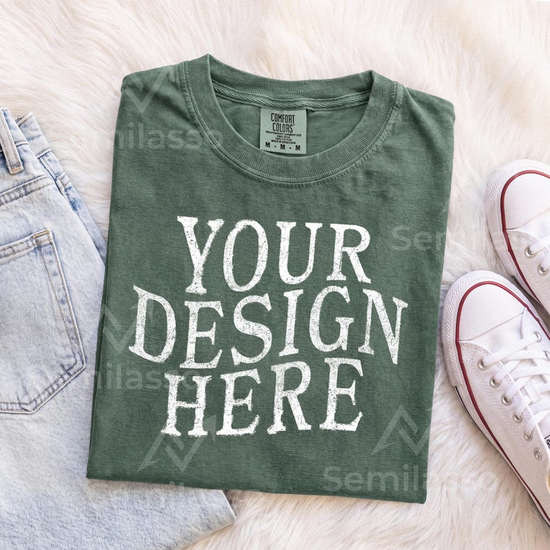 May include: A dark olive green crew neck t-shirt featuring the white text "YOUR DESIGN HERE" printed in a distressed, bold font on the front. The shirt has a visible interior neck label indicating a size medium and a classic short-sleeve construction.