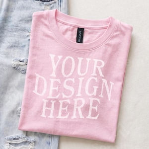 May include: A light pink short-sleeved t-shirt with a crew neckline and the white text YOUR DESIGN HERE printed on the front. The shirt features a black Gildan brand label at the inner neck and is positioned next to light blue distressed denim jeans.