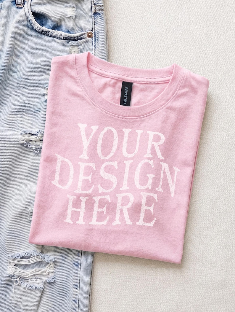 May include: A light pink short-sleeved t-shirt with a crew neckline and the white text YOUR DESIGN HERE printed on the front. The shirt features a black Gildan brand label at the inner neck and is positioned next to light blue distressed denim jeans.