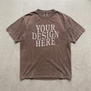 May include: A brown short-sleeve t-shirt with a crew neckline. The center of the shirt features the text "YOUR DESIGN HERE" printed in a distressed, off-white, serif font. The fabric has a muted, earthy tone with a soft, worn-in appearance.