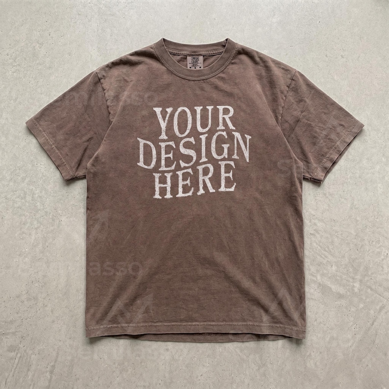 May include: A brown short-sleeve t-shirt with a crew neckline. The center of the shirt features the text "YOUR DESIGN HERE" printed in a distressed, off-white, serif font. The fabric has a muted, earthy tone with a soft, worn-in appearance.