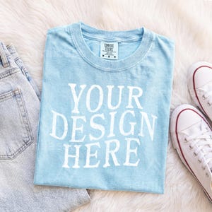May include: A light blue short-sleeve t-shirt with a crew neckline and the white text "YOUR DESIGN HERE" printed on the front. The shirt features a Comfort Colors brand label on the inside collar indicating a size medium.