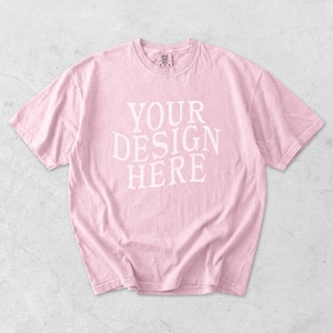 May include: A light pink t-shirt with the text "YOUR DESIGN HERE" in white block letters. The shirt has short sleeves and a crew neck. The fabric appears soft and slightly wrinkled.