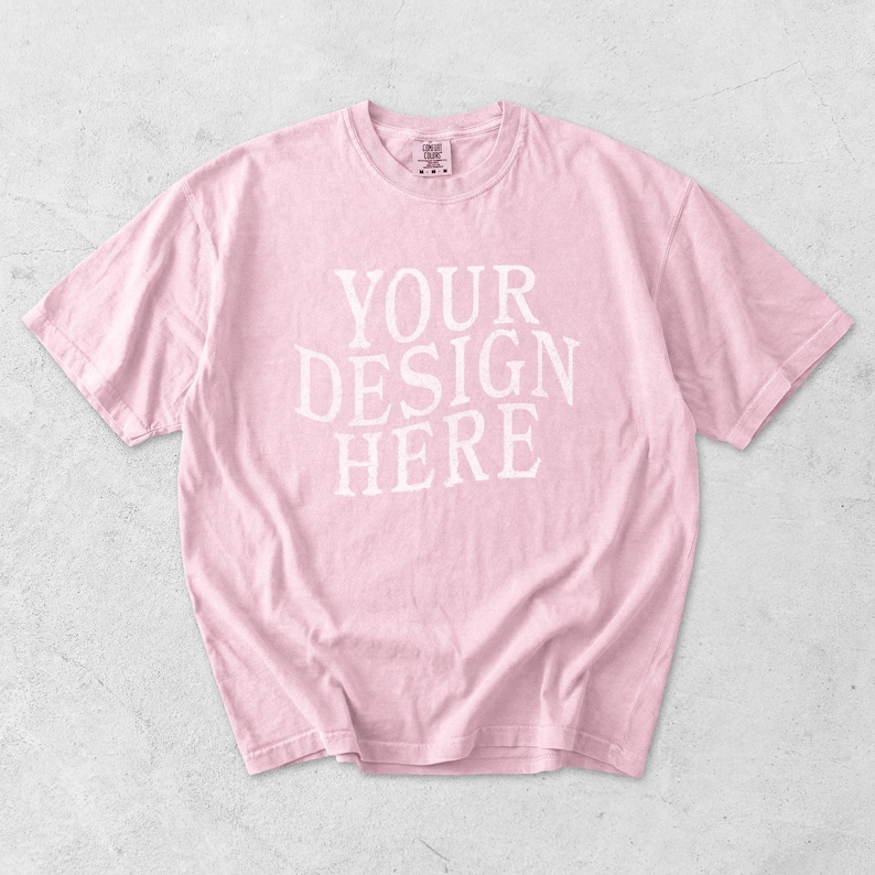 May include: A light pink t-shirt with the text "YOUR DESIGN HERE" in white block letters. The shirt has short sleeves and a crew neck. The fabric appears soft and slightly wrinkled.
