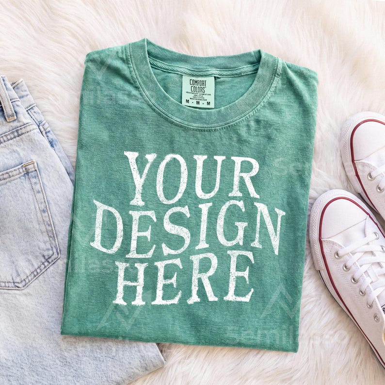 May include: A green short-sleeved t-shirt with a crew neckline and a white graphic print that reads "YOUR DESIGN HERE" in a bold, distressed font. The shirt features a size M tag and is displayed alongside light blue denim and white sneakers.