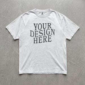 May include: A short-sleeve crew neck t-shirt in a light gray or off-white color. The center of the chest features dark gray, distressed-style lettering that reads YOUR DESIGN HERE in a bold, serif font. The garment has a standard fit.