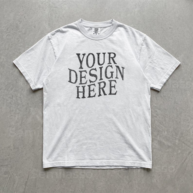 May include: A short-sleeve crew neck t-shirt in a light gray or off-white color. The center of the chest features dark gray, distressed-style lettering that reads YOUR DESIGN HERE in a bold, serif font. The garment has a standard fit.