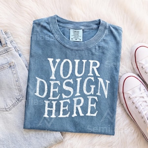 May include: A blue short-sleeved t-shirt with a crew neckline and a white graphic print that reads YOUR DESIGN HERE. The shirt features a tag at the inner collar indicating size M and is made of 100% cotton, as shown on the garment label.
