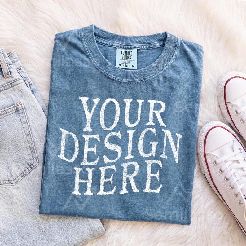 May include: A blue short-sleeved t-shirt with a crew neckline and a white graphic print that reads YOUR DESIGN HERE. The shirt features a tag at the inner collar indicating size M and is made of 100% cotton, as shown on the garment label.