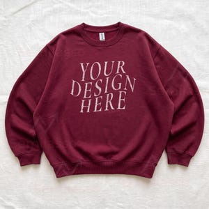 Puede incluir: Una sudadera de color granate con cuello redondo y manga larga que presenta el texto "YOUR DESIGN HERE" impreso en una fuente blanca desgastada en el centro del pecho. La prenda cuenta con cuello, pu&ntilde;os y dobladillo acanalados.