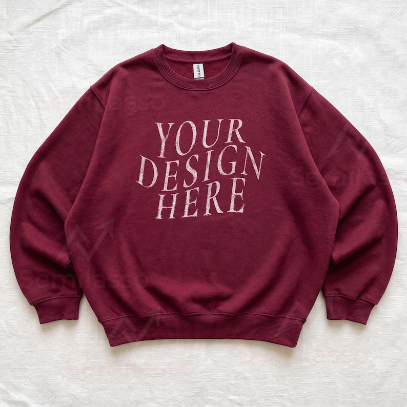 Puede incluir: Una sudadera de color granate con cuello redondo y manga larga que presenta el texto "YOUR DESIGN HERE" impreso en una fuente blanca desgastada en el centro del pecho. La prenda cuenta con cuello, pu&ntilde;os y dobladillo acanalados.