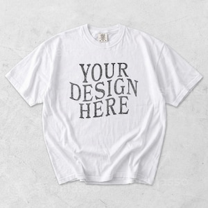 May include: White t-shirt featuring the text "YOUR DESIGN HERE" in a distressed, dark grey font. The shirt has a crew neck and short sleeves. The garment is displayed flat on a neutral background.