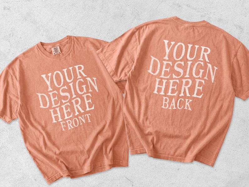 May include: Two coral-colored short-sleeve t-shirts displayed together. The left shirt features white text reading YOUR DESIGN HERE FRONT. The right shirt displays white text reading YOUR DESIGN HERE BACK. Both shirts have a crew neckline.