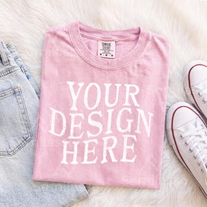 May include: A light pink short-sleeved t-shirt with the white text YOUR DESIGN HERE printed on the front. The shirt features a crew neckline and a Comfort Colors brand label on the inside collar indicating a size medium.
