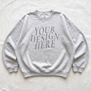 May include: A light gray heather crewneck sweatshirt featuring the dark gray text "YOUR DESIGN HERE" printed in a centered, slightly arched serif font on the chest. The garment has long sleeves, ribbed cuffs, and a ribbed neckline.