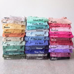 Puede incluir: Una colección de camisetas dobladas dispuestas en tres pilas verticales, que muestran una gran variedad de colores incluyendo blanco, amarillo, azul, verde, rosa, rojo, morado, gris y negro, cada una etiquetada con su nombre de color impreso.