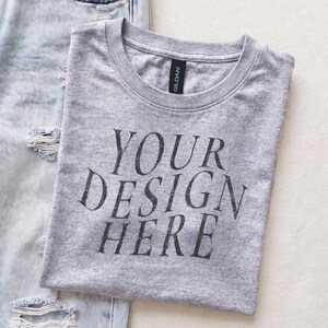 May include: A heather gray crew neck t-shirt with the black text "YOUR DESIGN HERE" printed on the front, positioned next to a pair of light blue distressed denim jeans with visible rips and frayed edges on the fabric surface.