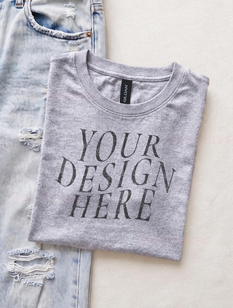 May include: A heather gray crew neck t-shirt with the black text "YOUR DESIGN HERE" printed on the front, positioned next to a pair of light blue distressed denim jeans with visible rips and frayed edges on the fabric surface.