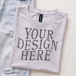 May include: A light gray short-sleeved crew neck t-shirt with the text "YOUR DESIGN HERE" printed in dark gray letters on the front. The shirt is positioned next to a pair of light blue distressed denim jeans with visible rips and frayed edges.