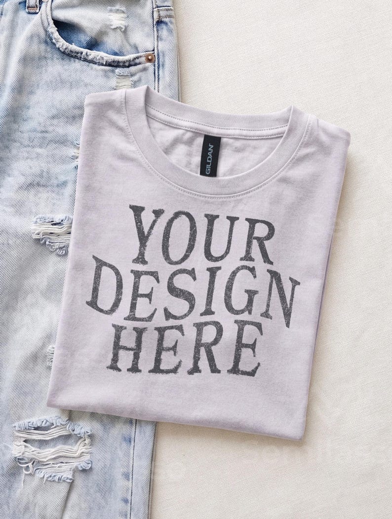 May include: A light gray short-sleeved crew neck t-shirt with the text "YOUR DESIGN HERE" printed in dark gray letters on the front. The shirt is positioned next to a pair of light blue distressed denim jeans with visible rips and frayed edges.