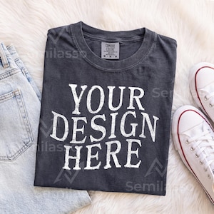 May include: A charcoal gray short-sleeve t-shirt with a crew neckline and a distressed white graphic print that reads YOUR DESIGN HERE. The shirt features a Comfort Colors brand label on the inside collar and is made of cotton fabric.